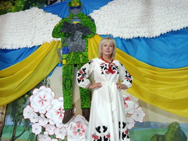 LaurenWishy stands in front of a colorful, layered backdrop with blue, yellow, and white fabric. She wears a white dress with red and black floral patterns, hands on hips. Behind her, a green, armored figure with wings and a helmet is centered. The figure has gray armor and holds a blue object. Large white and pink flowers are at the bottom, partially obscuring the figure's legs. The background has a painted, forest-like scene. She has blonde hair and a neutral expression.
