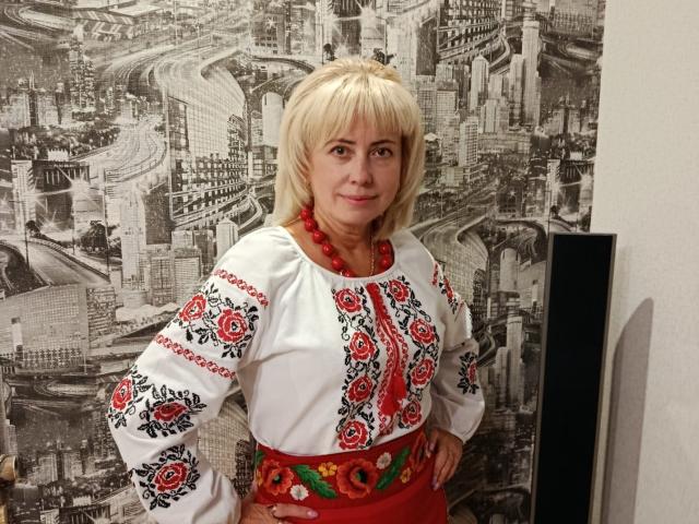 LaurenWishy stands in a room with black-and-white cityscape wallpaper. She has blonde hair and wears a white blouse with red floral embroidery on the sleeves and chest, a red beaded necklace, and a red floral skirt. Her hands are on her hips, and she looks at the camera with a slight smile. The blouse's embroidery is detailed with red and black patterns, and a red flower is centered on the waist. The overall style is traditional with a modern city backdrop.