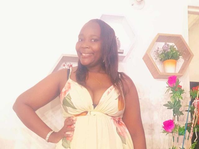 LustyEbonyMilf with dark skin, smiling, wearing a floral dress, showing her tits, standing with hand on hip, bracelet on wrist, and pink roses in the background.