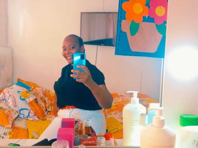 LustyEbonyMilf, bald, smiling, black shirt, white pants, holding phone, mirror selfie, messy bed, colorful blanket, toiletries on mirror, blue flower wall art, bright room.