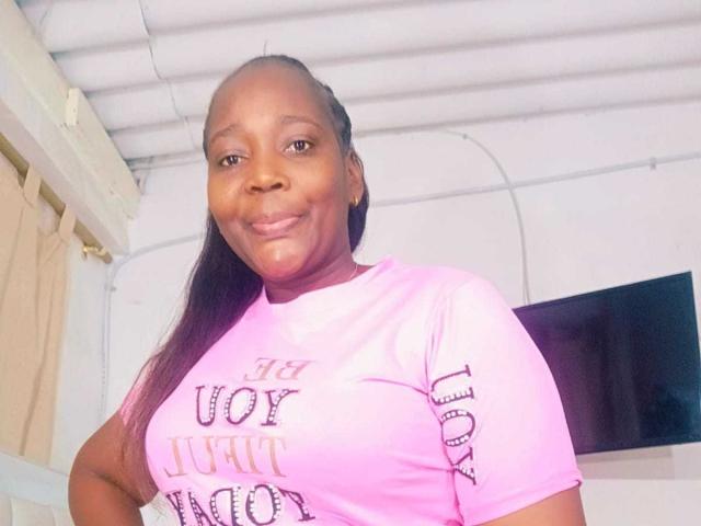 LustyEbonyMilf with dark brown skin, long straight black hair, pink t-shirt, and small tits, smiling slightly, standing indoors, wearing small hoop earrings, looking at camera.