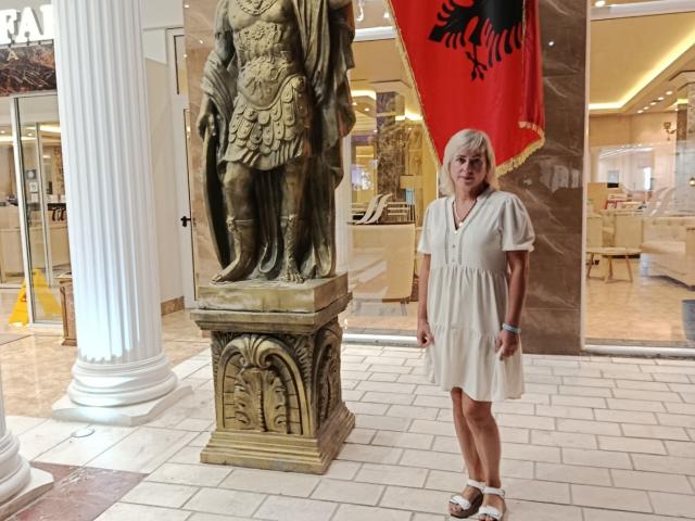 LaurenWishy stands next to a bronze statue of a Roman soldier. She's wearing a short, light beige dress with white sandals. Her white hair contrasts with the dark statue. A red flag with a black emblem is in the background. The floor is tiled in white and beige. LaurenWishy has a blue wristband on her right wrist. The setting is a well-lit, indoor space.