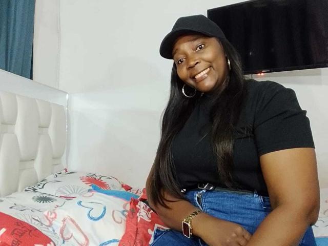 LustyEbonyMilf, dark-skinned, curvy, with long black hair, wearing black cap, black shirt, blue jeans, smiling, sitting on bed, white headboard, colorful bedspread, hoop earrings, gold watch.