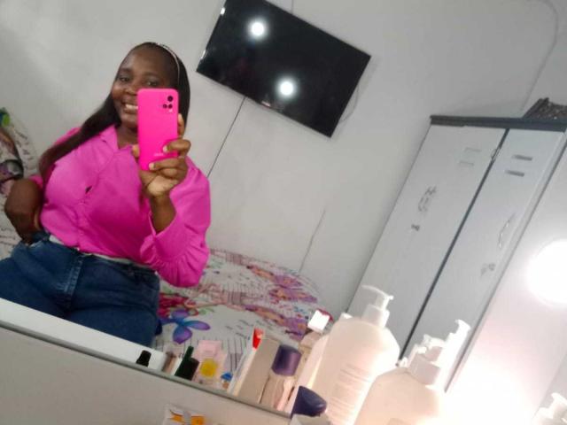 LustyEbonyMilf with dark skin, long straight hair, pink shirt, high-waisted blue jeans, holding pink phone, mirror selfie, white wall, TV, and various bottles on countertop.
