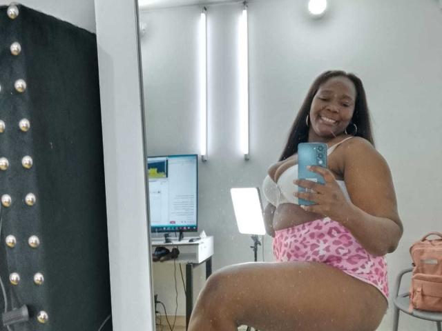 LustyEbonyMilf with dark skin, long brown hair, smiling, wearing white bra, pink leopard print panties, holding blue phone, taking mirror selfie, showing big ass, in room with computer.