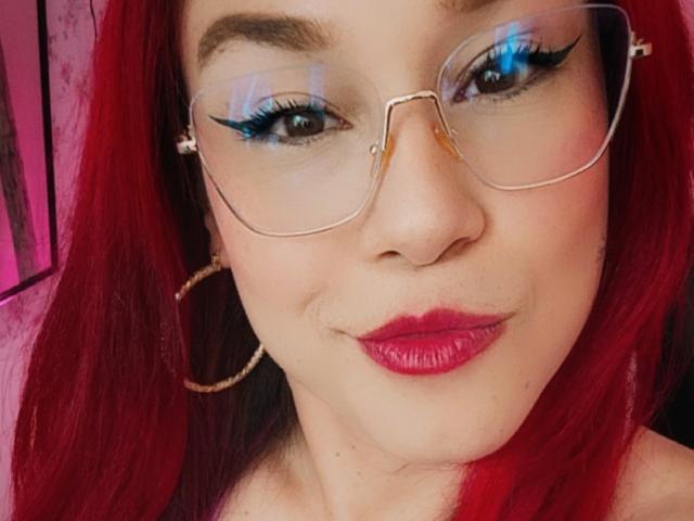 KimBurton with red hair, wearing glasses, blue eyeliner, pink lipstick, and gold hoop earrings. Her eyes are half-open, giving a subtle, confident smile.