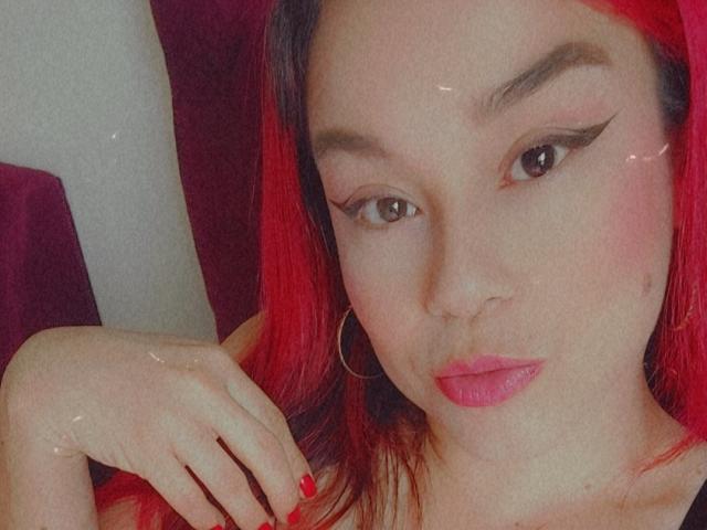 KimBurton with red hair, winged eyeliner, pink lipstick, hoop earrings, and red nails, looking at camera with a slight pout, hand near face.