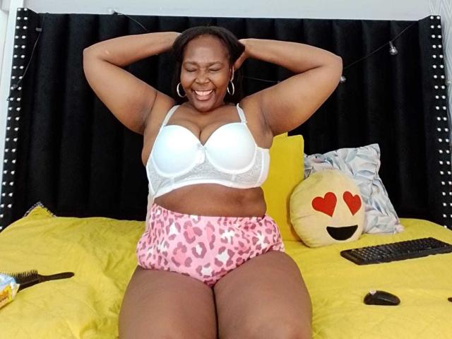 LustyEbonyMilf, curvy with dark skin, smiling, hands behind head, white bra, pink leopard print panties, sitting on yellow bed, black curtains, happy expression, thick thighs, small hoop earrings.
