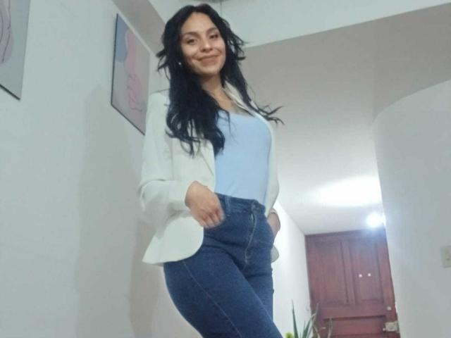Antonia Honey hat langes, schwarzes Haar, ein hellblaues Tanktop, einen weißen Blazer und blaue Jeans. Sie steht mit einem leichten Lächeln, die Hände in den Taschen, und sieht niedlich aus..