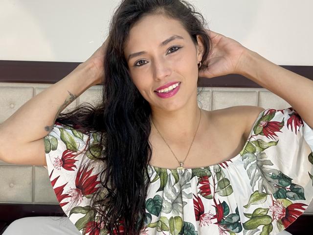 Kimberly Oobos tiene el pelo largo y negro, ojos marrones y una sonrisa. Lleva un vestido con estampado floral, tiene las manos en el pelo, un collar y pintalabios rosa. Los senos son pequeños y se ven debajo del vestido..