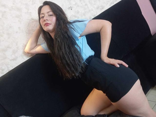 ScarlettSweetts with long black hair, light blue shirt, black shorts, leaning on black couch, hand on hip, red lipstick, confident expression, knee on couch, pale skin.