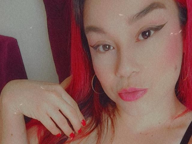 KimBurton has red hair, pink lips, and wears black eyeliner with winged tips. Her hand with red nails rests near her face. She's wearing a black top.