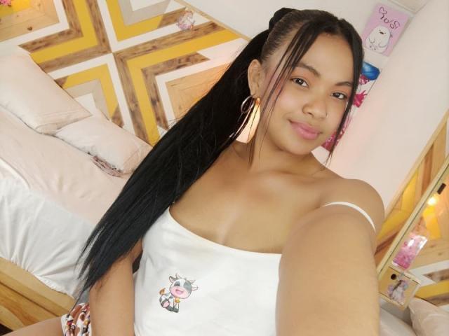 Solene Fontaine, con cabello negro largo, vestida con una camiseta blanca que deja ver pequeños senos, sonriendo, con grandes aretes dorados, tomando un selfie en una habitación muy decorada..