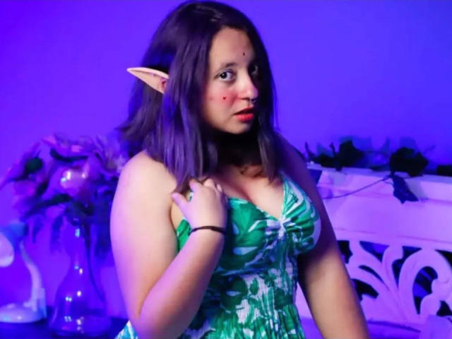 CristinaSmit with elf ears, purple hair, green leaf-pattern dress, holding her boob, purple blue light, black choker, red zit on her cheek, looking at camera.