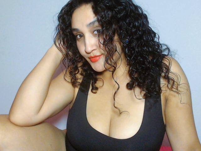 JasminePark with curly black hair, wearing a black tank top, showing her large tits, sitting with a hand in her hair, and red lipstick.