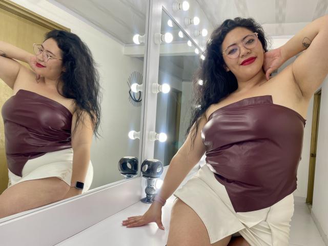 ErikaFox with curly black hair, red lipstick, glasses, brown strapless top, white skirt, mirror reflection, arm raised, smiling, armpit visible, black wristwatch, light bulbs in background.