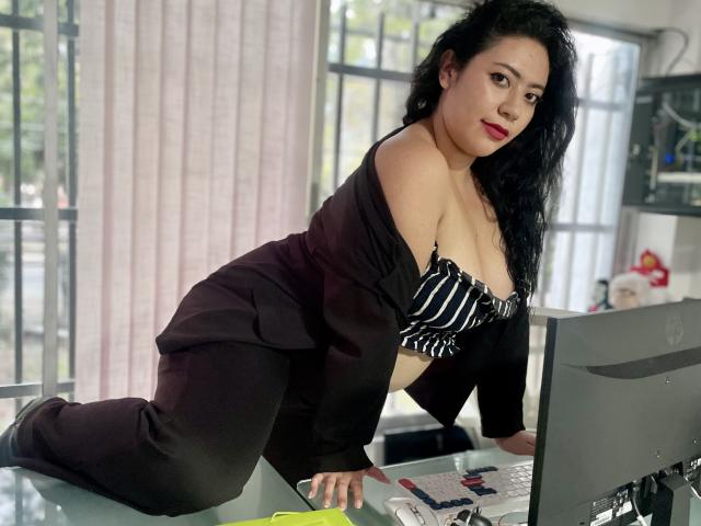 ErikaFox with black hair, red lipstick, and medium tits, wearing a black open shirt and striped bra, kneeling on desk, leaning on computer, in office setting.