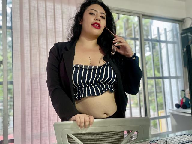 ErikaFox with curly black hair, red lipstick, black blazer, striped crop top, holding glasses, standing by desk, showing belly, confident, sexy.