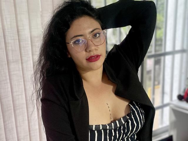 ErikaFox has wavy black hair, wears glasses, a black blazer, and a striped top showing her tits. She's looking at the camera with red lipstick.