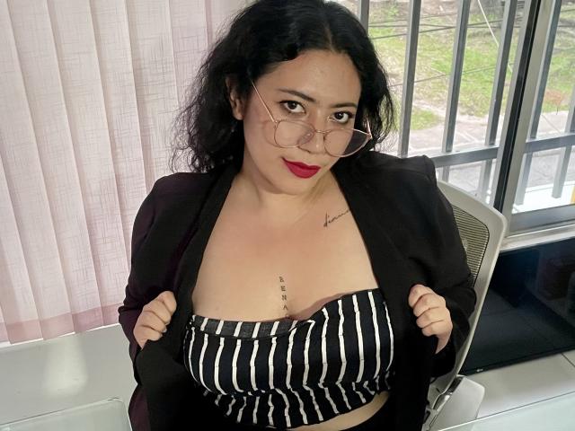ErikaFox with curly black hair, red lipstick, and glasses, wearing a black blazer and striped tube top, exposing her boobs, has a tattoo under her right breast.