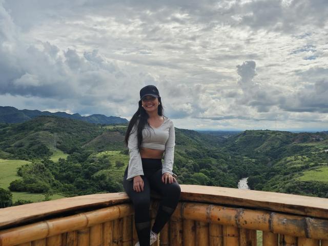 SophieJones69 with long black hair, smiling, wearing a white crop top, black cap, and black pants, sitting on a bamboo railing, with green hills and cloudy sky behind.