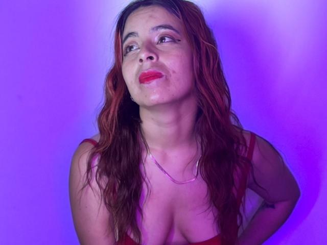 MeghanStorm69 with wet red hair, red lipstick, and a slight smile, wearing a red dress, showing her tits, against a purple background, looking upward with a subtle gaze.