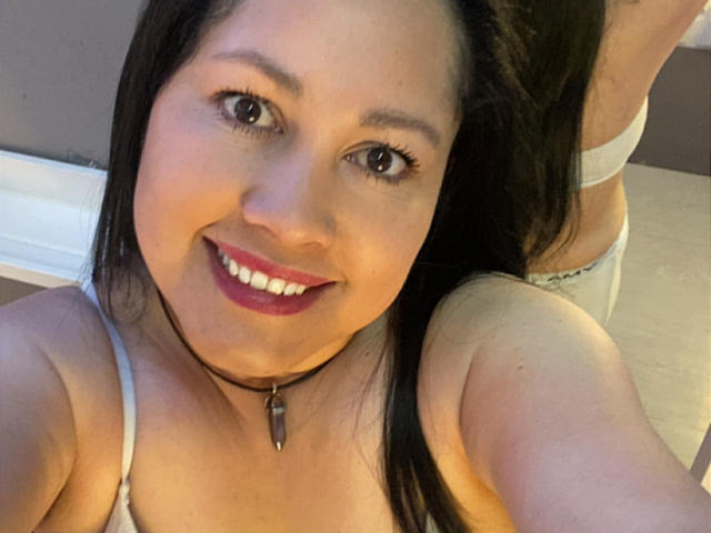 Mariana Smitt, com olhos castanhos, sorrindo, usando um colar preto com um pingente de bala, sutiã branco e calcinha cinza, seus seios e nádegas visíveis ao fundo..