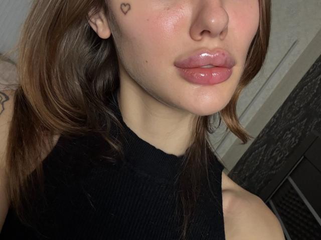 SensualMila's face with glossy pink lips, a small heart tattoo near her right eye, brown hair, and a black sleeveless top, showing a tattoo on her left arm.