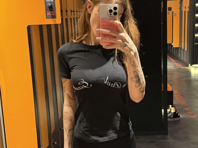 SensualMila taking a mirror selfie in a locker room, wearing a black t-shirt that slightly shows her tits, with tattoos on her arms and holding a pink phone.