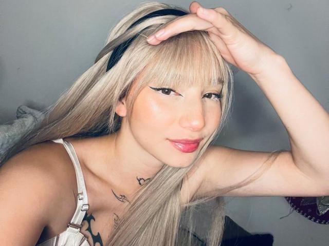 NicoleeCam with blonde hair, black headband, and pink lipstick, touching her forehead, wearing a white top, showing a small tattoo under her collarbone, smiling slightly.