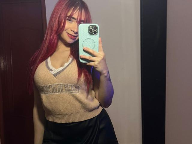 MelannyTaylorr with red hair, smiling, wearing a beige V-neck sweater and black satin pants, holding a blue phone, showing her tits in a mirror selfie.