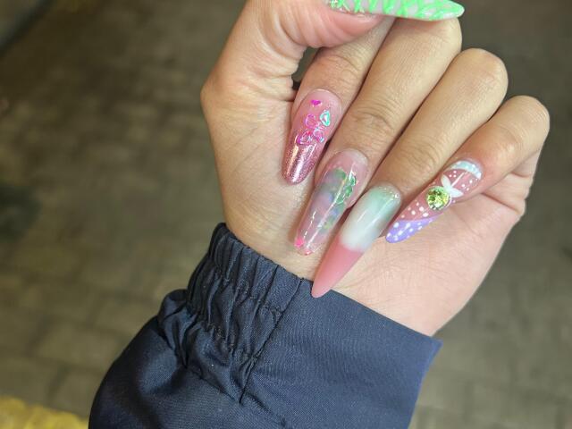 JulietaFoster69's hand with long, glittery nails. Nails feature pink, green, and purple designs, with glittery and clear patterns. JulietaFoster69 wears a black shirt.