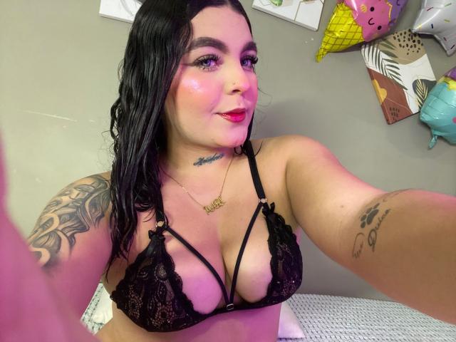 MelodyBlossom trägt einen schwarzen Spitzen-BH, der ihre großen Brüste zeigt, hat Tattoos an den Armen, trägt rosigen Rouge und hat schwarzes Haar. Sie macht ein Selfie, trägt eine goldene Halskette und hat rosigen Lippenstift..