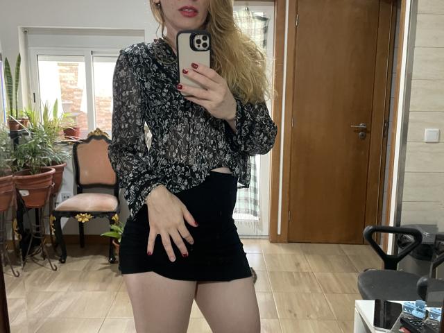 AnaliceColin stands in a room, taking a mirror selfie with a smartphone. She has long, wavy blonde hair and wears a black floral blouse with long sleeves and a black, high-waisted mini skirt. Her right hand is placed on her skirt, and her nails are painted red. The room has wooden floors, a potted plant, and a wooden chair with a floral cushion. The door in the background is wooden, and there's a small black chair on the right. The image is clear and focused.