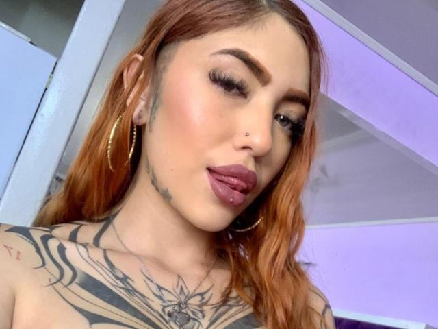 AntonellaBrith has orange hair, gold hoop earrings, tattoos, and a nose ring. She's sticking out her tongue, with glossy pink lips and a slight blush. Boobs visible.
