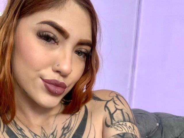 AntonellaBrith has light skin and shoulder-length orange hair. She has a nose piercing, a lip piercing, and thick, dark eyebrows. Her eyes are brown with long, dark eyelashes. She's wearing dark purple lipstick. AntonellaBrith's tattoos are visible on her shoulders and chest, featuring black, intricate designs. She's looking at the camera with a slight, confident smile. The background is a plain, light purple wall. The image has a casual, selfie-style composition.