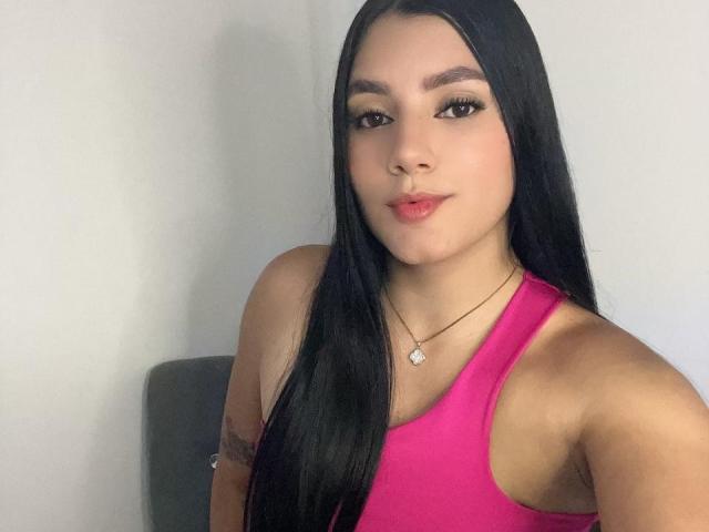 CristalAngels with long black hair, pink tank top, and a small tattoo on her right arm, wearing a necklace with a heart-shaped pendant, looking at the camera.