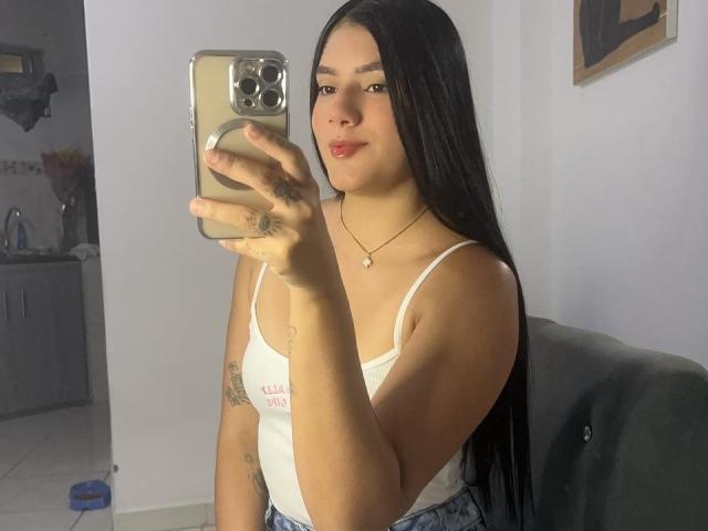 CristalAngels with long black hair, wearing a white tank top, holding a phone, showing tattoos on her hand and arm, taking a mirror selfie, showing her tits.