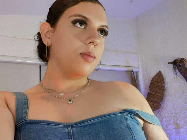CatleyaNoire, con i capelli castani raccolti in uno chignon, indossa un abito di jeans blu con una spalla scoperta, orecchini a cerchio e una collana, e guarda di lato con un sorriso appena accennato..