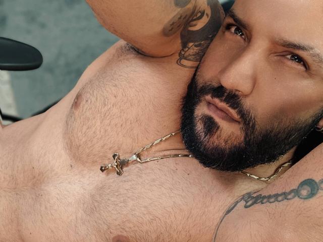 BardoCkroy69 has a hairy chest with a cross necklace, tattoos on arms, and a beard, lying down, looking at the camera, showing his chest and arm tattoos.
