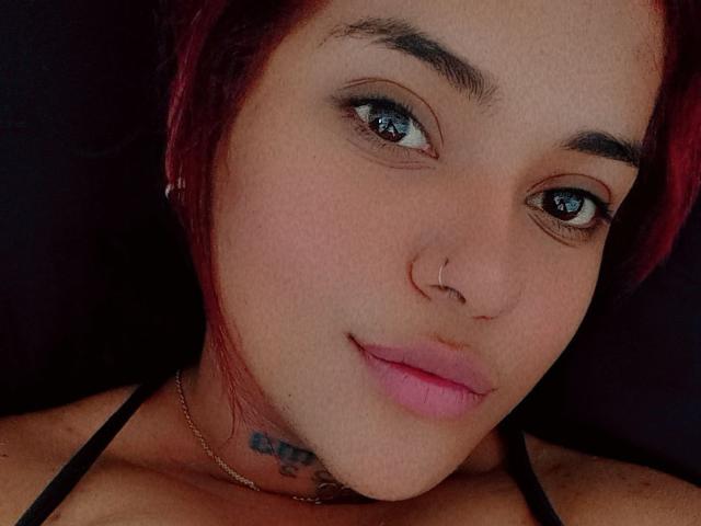 NaomyNoa hat rote Haare, einen Nasenring und rosafarbene Lippen mit einem leichten Lächeln. Sie trägt ein schwarzes Top mit Halter und zeigt dabei ein kleines Tattoo unter ihrem Kinn..
