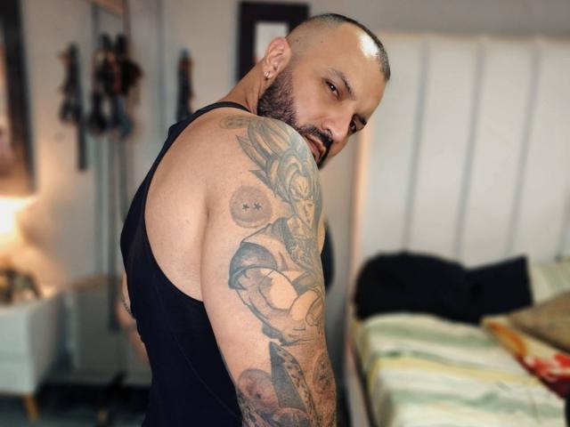 BardoCkroy69, bald with a beard, wearing a black tank top, showing tattooed arms, looking back with a serious expression, in a bedroom with a bed and lamp.