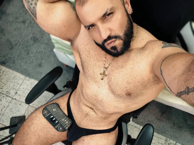 BardoCkroy69, muscular, hairy, bearded, wearing black jockstrap with silver emblem, gold necklace, tattoos, seated, arms raised, looking up, semi-erect, tan skin, confident, chair, overhead shot.