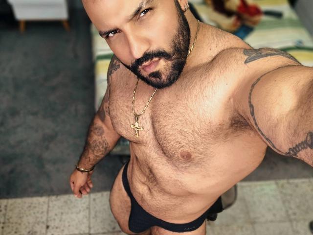 BardoCkroy69, a hairy, muscular man with tattoos, black beard, and gold cross necklace, wears a black jockstrap, looking up with a serious expression.