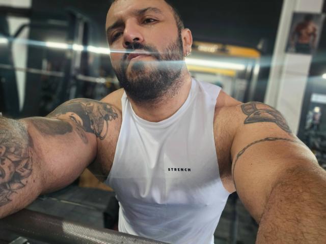 Tattooed BardoCkroy69 with a beard, white tank top, and a bulge in the gym, holding a phone, taking a selfie. Visible tattoos on arms. Bearded, muscular, bald.