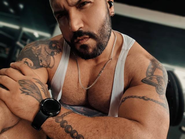 BardoCkroy69 with a beefy, hairy chest, white tank top, tattoos, black wristwatch, chain necklace, and serious expression. His arms are crossed, showing a muscular, hairy body.