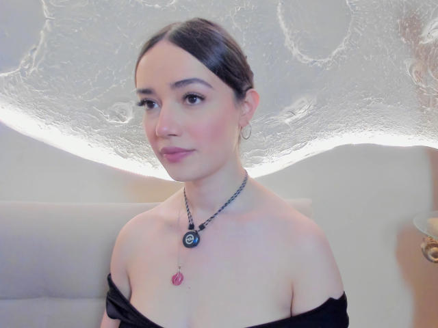 MariaLoft, com cabelo escuro, pele clara e blush rosa nas bochechas, vestindo uma blusa preta com um ombro descoberto e um colar com uma pedra rosa e um anel azul..