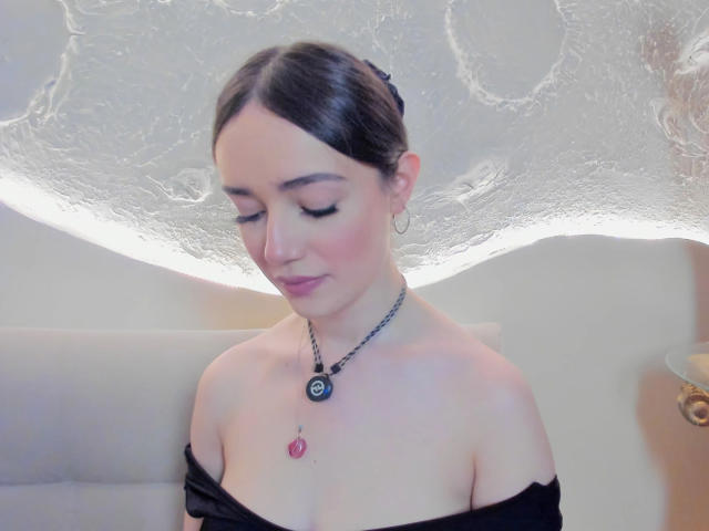 MariaLoft, con cabello oscuro, ojos cerrados, blusa negra con un hombro descubierto, pechos pequeños, rubor rosado, collar con dije negro y rosa, aretes de aro plateados, mirando hacia abajo, en luz suave..