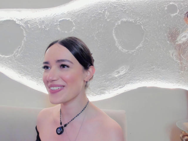 MariaLoft, avec ses cheveux foncés attachés en chignon, souriante, portant un haut noir à une épaule, un collier avec un pendentif circulaire et des boucles d'oreilles cerceaux, sur un fond blanc texturé, évoquant des bulles..