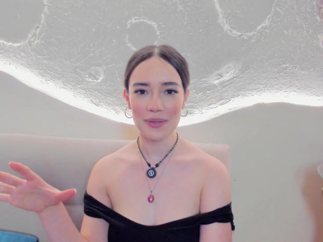 MariaLoft, met donker haar in een knot, draagt een zwarte top met een open schouder, oorringen en een ketting met een rode steen, lachend en haar linkerhand omhoog houdend..